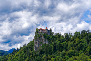 Fototapeta na wymiar Castle on top of rock with waving Slovenian flag at lakeshore of Slovenian Lake Bled on a blue cloudy summer day. Photo taken August 8th, 2023, Bled, Slovenia.
