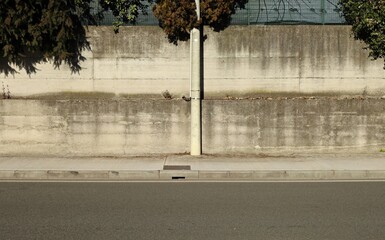 Cement pole on sidewalk with precast concrete wall on behind. Branches of tree on top, urban street...