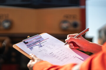A service technician is checking on heavy machine maintenance checklist, with an ancient train locomotive head as blurred background. Transportation industrial working scene, selective focus at hand.