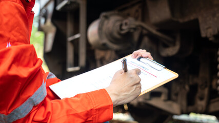 	
Action of a mechanic engineer is checking on heavy machine checklist form to verify the quality of maintenance service, with train locomotive part as blurred background. Industrial working scene.