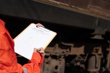 Action of a mechanic engineer is checking on heavy machine checklist form to verify the quality of maintenance service, with train locomotive part as blurred background. Industrial working scene.