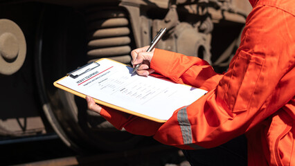 Action of a mechanic engineer is checking on heavy machine checklist form to verify the quality of maintenance service, with train locomotive part as blurred background. Industrial working scene.