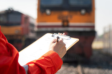A service technician is checking on heavy machine maintenance checklist, with an ancient train locomotive head as blurred background. Transportation industrial working scene, selective focus at hand.