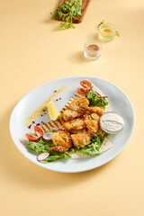 Tempura shrimp with tartar sauce on a bed of lettuce, presented on a white plate with lemon and tomato accents, against a soft yellow backdrop