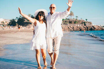 Happy senior couple walking barefoot on the seashore at sunset enjoying vacation and retirement,...