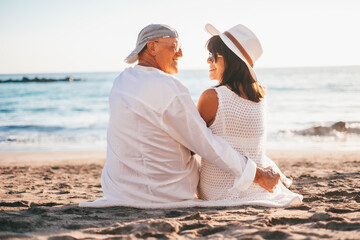 Rear view of romantic senior couple sitting on the beach face the sea enjoying vacation and retirement, two elderly people expressing love and tenderness