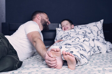 Bare feet of a child. Dad tickles his child's feet sticking out from under the blanket. Laughter and joy of father and son. - obrazy, fototapety, plakaty