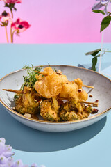 Elegant tempura vegetables on skewers with nut sauce amidst a surreal pastel floral backdrop