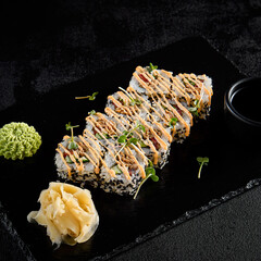 Sushi roll with vegetables and fried salmon inside, sesame seeds outside, and spicy sauce, presented on a black slate with soy sauce, wasabi, and ginger