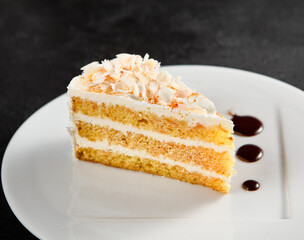 A slice of white chocolate and coconut cake on a white plate with dark syrup, against a textured black backdrop