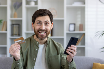 Excited buyer with debit card and mobile phone in pleasant shock from online shopping from home. Amazed male customer getting massive cash back and extra bonuses after making order with shipping.