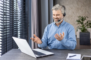 Elderly grey-haired man in denim shirt sitting at domestic workspace with laptop and having distant...