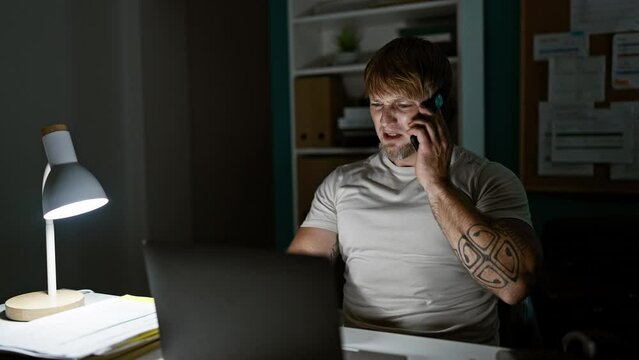 A concentrated young man with a beard working late on his laptop in a dimly lit office while talking on the phone.