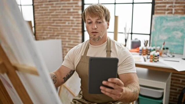 A focused caucasian man with a beard using a tablet in a creative studio filled with paints and art supplies.