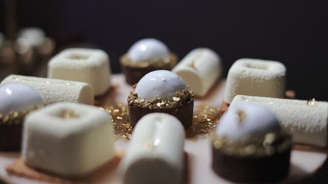 Assortment of sweet smooth snacks in light colours decorated with golden foil flakes on festive candy bar. Collection of creamy desserts arranged in circle on board for birthday party reception.