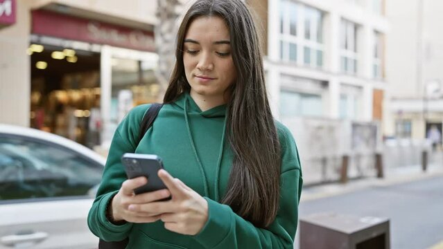 Young hispanic woman using smartphone on busy city street backdrop, portraying an urban teen's lifestyle.