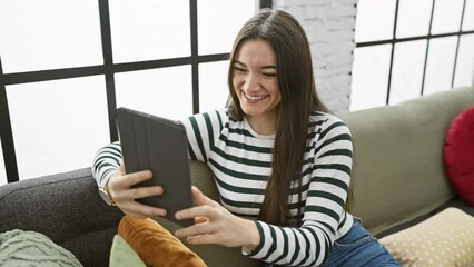 Hispanic woman reading tablet on sofa in a cozy living room, portraying relaxed indoor leisure time.