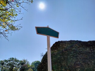 In the picture is the atmosphere in the late morning of one day with the sky, branches, sunlight, the old city wall, and a blue sign with the handle of a white sign standing near the old wall.