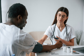 Fototapeta premium Sitting by a table. Female doctor measures blood pressure of a man
