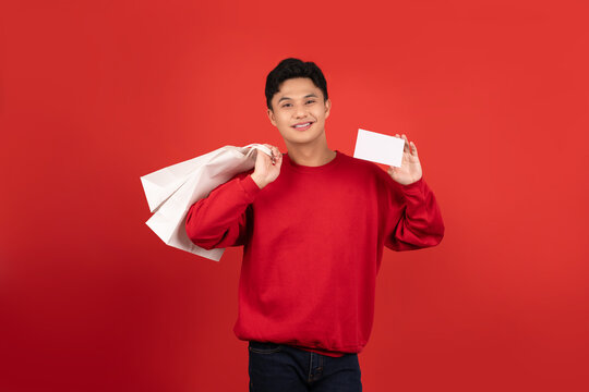 Young Asian man wearing a red sweater is holding a credit card and shopping bags on a red background.