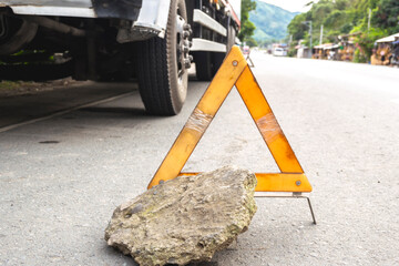 A weathered emergency warning triangle supported by a rock on a roadside, with a blurry truck in...