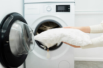 Woman holding clean white pillow in front of the drum of washing machine in laundry room. Washing pillows - obrazy, fototapety, plakaty