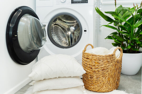 Laundry basket and pile of white pillows in front of the opened washing machine in laundry room. Housekeeping