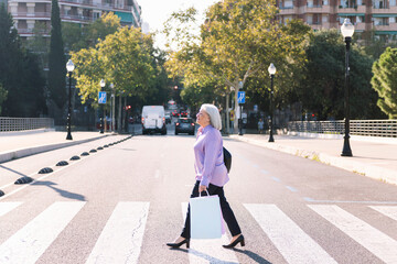 senior woman crossing a city street at a pedestrian crossing with shopping bags in her hand, concept of elderly people leisure and active lifestyle © Raul Mellado
