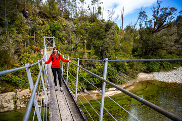 hiker girl crossing a hanging bridge over pororari river, west coast of new zealand south island, bushwalking through beautiful temperate rainforest