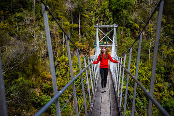 Fototapeta na wymiar hiker girl crossing a hanging bridge over pororari river, west coast of new zealand south island, bushwalking through beautiful temperate rainforest