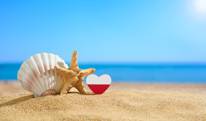 Poland flag in the shape of a heart and shells on a sandy beach.