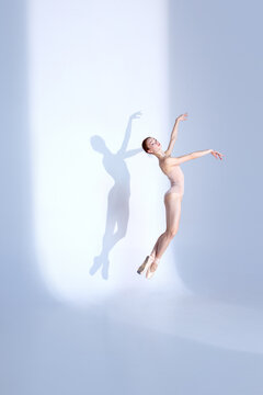 Graceful Ballet. Portrait of young woman dances in pink sport swimsuit and pointe against white studio backdrop, her shadow mirroring her grace. Concept of elegance, athleticism, ballet.