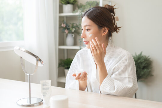 Facial beauty skin care, smile of pretty asian young woman in bathrobe looking at mirror, hand applying moisturizer lotion on her face, holding jar of skin cream before makeup cosmetic routine at home - Powered by Adobe