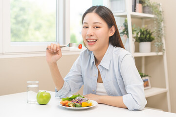 Diet concept, happy asian young woman hand use a fork to prick tomato, fresh vegetable or green salad, eat nutrition food  on table at home, low fat to good body. Girl getting weight loss for healthy.