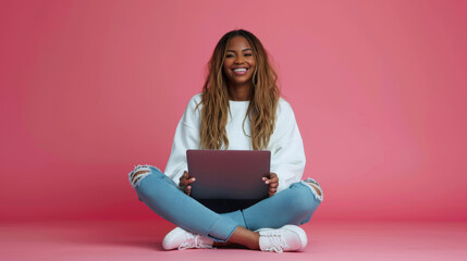 cheerful young woman sitting with her legs crossed, holding a laptop on her lap, wearing a white sweatshirt and ripped blue jeans, against a coral pink background