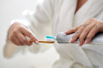 Hands of young woman in soft white bathrobe squeezing out toothpaste on toothbrush while standing...