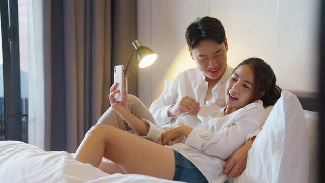Asia people young adult man woman relax hug smile look camera take photo record video post to social media at hotel cozy bed enjoy date day. Happy asian lover wife husband just married sweet newlywed