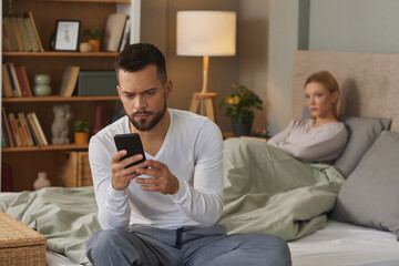 Man with smartphone in bedroom, upset woman in the background