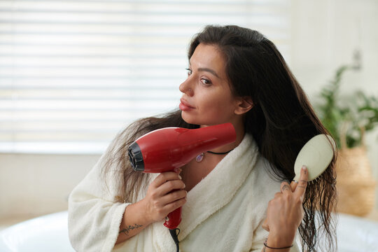 Brunette girl in white bathrobe brushing and drying her long dark hair with hair dryer after washing them while standing in bathroom