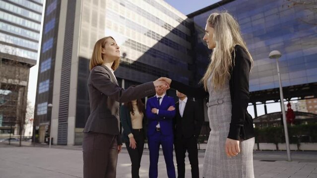 two executive business women bosses shaking hands to agree on a deal with employees looking behind coworking meeting