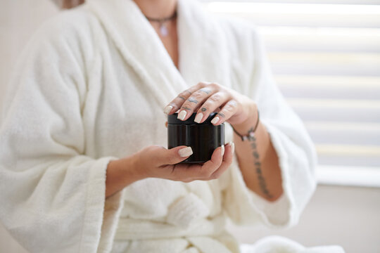 Hands of young woman in soft white bathrobe turning lid of black plastic jar with moisturizing cream or body scrub before beautycare procedure
