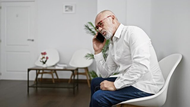 A senior man in a white shirt and glasses talks on the phone while sitting in a modern waiting room with white chairs and a decorative plant.