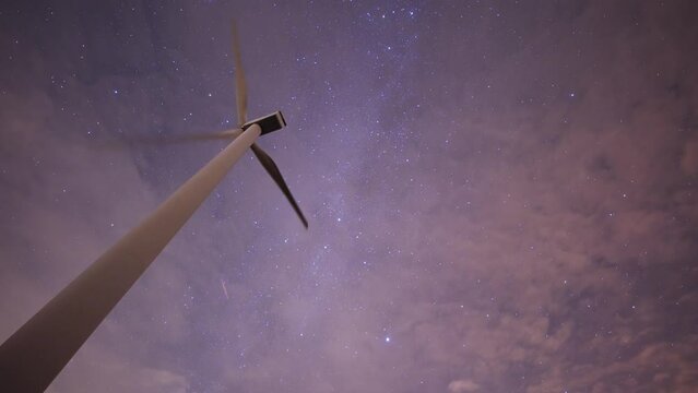 Bright stars and satellites in the cloudy night sky. A slowly rotating wind turbine in the foreground. A timelapse video.