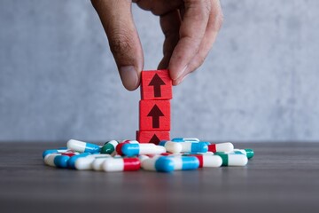 A red wooden block with upward-pointing arrow on a pile of colorful pills. Rising costs of...