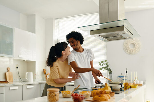 Young cheerful man looking at his wife while standing by electric stove in the kitchen and preparing something tasty for breakfast - Powered by Adobe