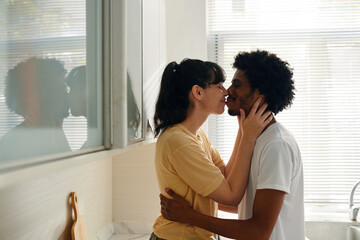 Young affectionate intercultural husband and wife in casualwear kissing each other while woman...