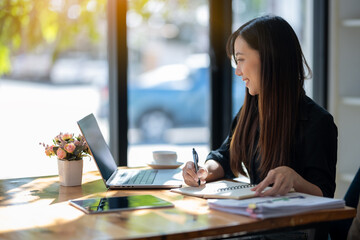 Obraz premium Attentive businesswoman jotting down notes while working on a project with her laptop at a sunny table..
