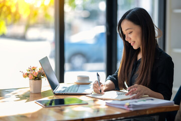 Obraz premium Attentive businesswoman jotting down notes while working on a project with her laptop at a sunny table..
