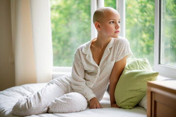 Obraz na płótnie Canvas Young bald girl wearing white pajama in hospital room sitting in bed and looking out the window.