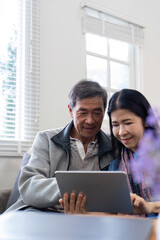 Happy elderly asian couple using tablet sit on sofa doing ecommerce shopping online on website and buying insurance browsing at home